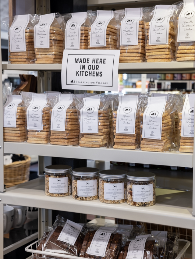 Shelves display neatly stacked bags of cookies and jars of snacks with white labels. A sign on the shelf reads, “MADE HERE IN OUR KITCHENS.” The setup is tidy and inviting in a specialty food store. Fearrington Village